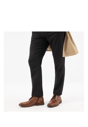 Ein Mann trägt schwarze Hosen, braune Leder-Schnürstiefel und hält einen beigen Mantel vor einem weißen Hintergrund.