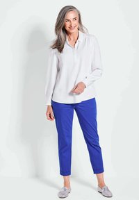 Weiße Bluse mit langen Ärmeln und Kragen, kombiniert mit blauen Slim-Fit-Hosen und grauen Slippern, vor einem neutralen Hintergrund.