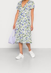 Robe midi florale avec un fond blanc, ornée de fleurs bleues et de feuilles vertes, à manches courtes bouffantes et décolleté en V profond. Portée avec des baskets blanches.
