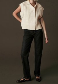 Gilet en tricot couleur crème avec texture côtelée, manches courtes et col à revers, associé à un jean noir droit et des ballerines pointues.