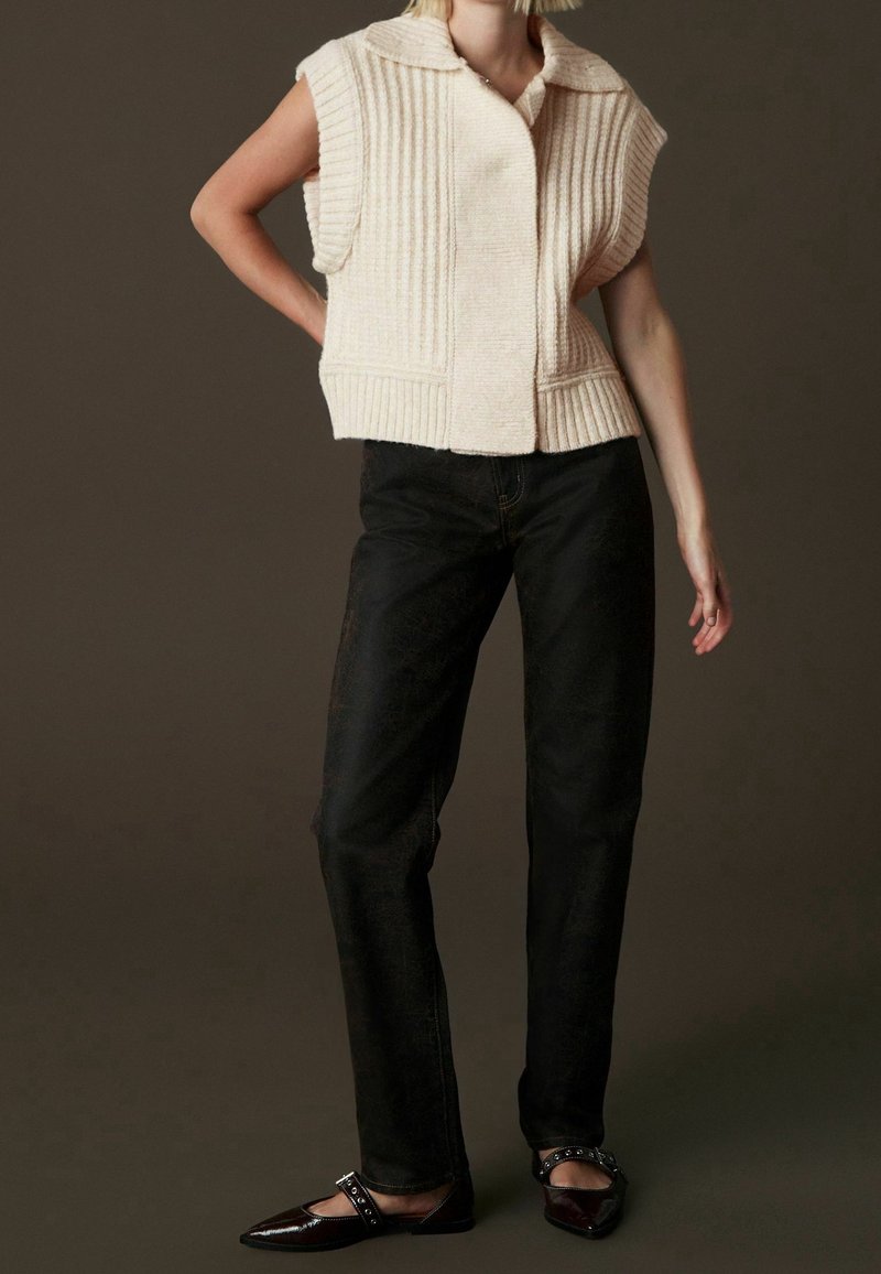 Gilet en tricot couleur crème avec texture côtelée, manches courtes et col à revers, associé à un jean noir droit et des ballerines pointues.