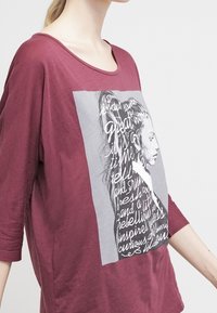 Maroon oversized t-shirt with a round neckline and three-quarter sleeves. Features a graphic print of a profile and text in black and white.