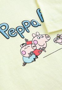 Vaaleankeltainen puuvillapaita, jossa on sarjakuvapossuja. Teksti kuuluu "Peppa Pig". Hahmoilla on vaaleanpunaiset kasvot, yksinkertaiset vaatteet ja kasvonpiirteet.