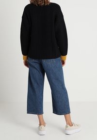 Czarny, dzianinowy, oversize sweter o prążkowanej strukturze z musztardowymi mankietami, w połączeniu z szerokimi, niebieskimi dżinsami i beżowymi sneakersami.