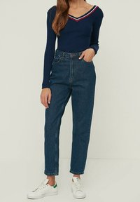 Ciemnoniebieski, prążkowany longsleeve z dekoltem w kształcie V i czerwonymi akcentami, w połączeniu z jasnoniebieskimi dżinsami typu high-waisted i białymi sneakersami.