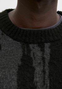 Nahaufnahme eines dunkelhäutigen Halses, der einen schwarzen gerippten Rundhalsausschnitt über einem grauen Hemd trägt.