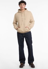 Felpa beige con cappuccio, tasca frontale, cordoncini e logo discreto. Abbinata a jeans dritti blu scuro e scarpe marroni.