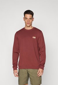Maroon langermet genser med ribbestrikkede mansjetter og nederst. Har en liten merkeetikett på venstre bryst. Glatt, bomullsblandingsstoff.