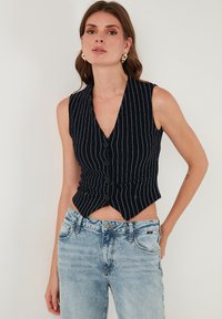 Námornícky pruhovaný bezrukávový vest s hlbokým výstrihom do V, s prednými gombíkmi, zúženým pásom a skráteným dizajnom, kombinovaný so svetlými džínsy.