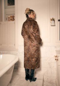 Manteau en fausse fourrure à imprimé léopard, long et ample, avec des bottines noires brillantes à la cheville. Texture lisse sur fond de salle de bain en marbre.