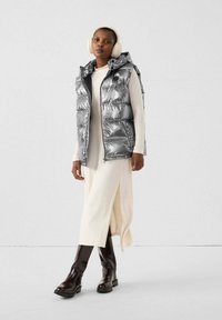 Gilet imbottito argento con cappuccio, abbinato a un vestito color crema con spacco laterale. La modella indossa stivali alti fino al ginocchio color marrone scuro e paraorecchie crema.