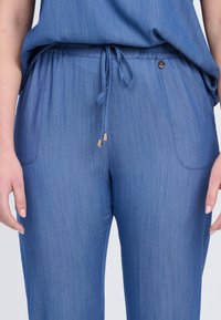 Helle blaue, taillierte Hose mit elastischem Bund, Kordelzugverschluss und goldenen Hardware-Akzenten. Glatte Textur mit dezenten vertikalen Streifen.