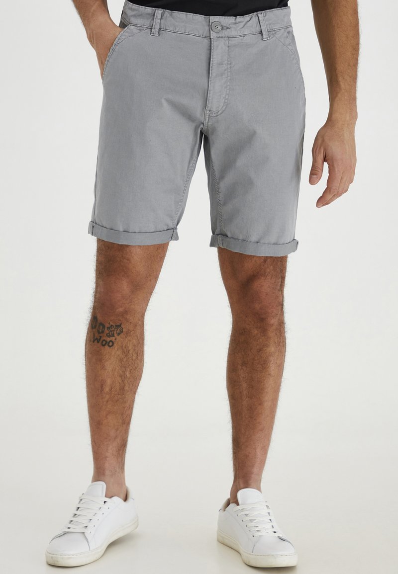Shorts en coton gris avec une fermeture à bouton, revers, poches latérales et un tatouage visible sur la jambe, assortis à des baskets blanches.