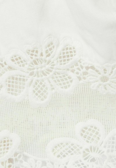 Tissu blanc avec une bordure en dentelle florale, doté d'un motif texturé et d'une broderie complexe, superposé sur un fond lisse et transparent.
