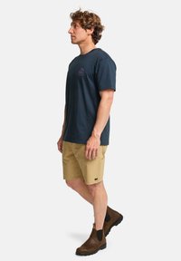 Camiseta de algodón azul marino con mangas cortas y un pequeño logo púrpura, combinada con pantalones cortos beige y botas de tobillo marrones. Diseño simple y casual.