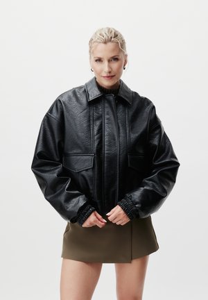 Blond kvinne i en svart oversized skinnjakke og olivengrønn miniskjørt som står foran en ensfarget hvit bakgrunn.