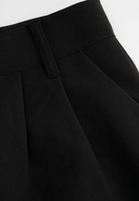 Czarna spódnica z wysokim stanem, z pętlami na pasek i plisowanymi detalami na jasnym tle.