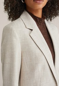 Beežikas tekstuuriga bleiser sisselõigatud kraega, millel on õrn ruuduline muster ja struktureeritud õlad, kihiliselt pruuni soonikuga topi kohal.