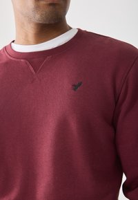 Sudadera burdeos con cuello redondo, que presenta un pequeño logo negro bordado en el pecho y una tela suave y texturizada.