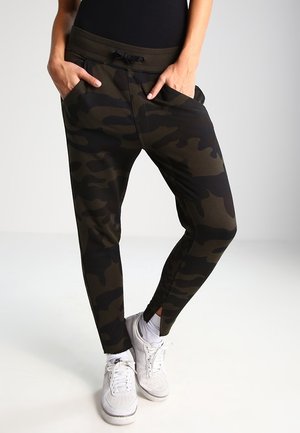 Personne portant un pantalon de jogging camouflage vert foncé et noir avec des poches, des chaussettes blanches et des baskets blanches Nike, debout les mains dans les poches.