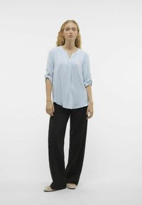 Lyseblå, tekstureret bluse med lange ærmer og V-udskæring; bæres med løse sorte bukser og lyse sko. Simpel, afslappet design.