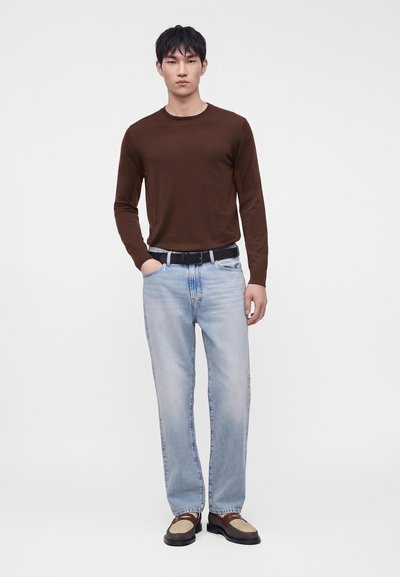 Homme portant une chemise marron à manches longues, un jean bleu clair avec une ceinture noire, et des chaussures bicolores marron et beige, debout devant un fond uni.