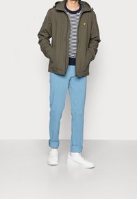 Person wearing an olive green jacket, navy and white striped shirt, light blue pants with cuffs, and white sneakers standing on a plain floor.