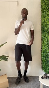 T-shirt bianco, pantaloni corti neri, calze nere e scarpe. Ambientazione interna con sfondo di piante verdi, scatola beige e una macchina fotografica su un tavolo.