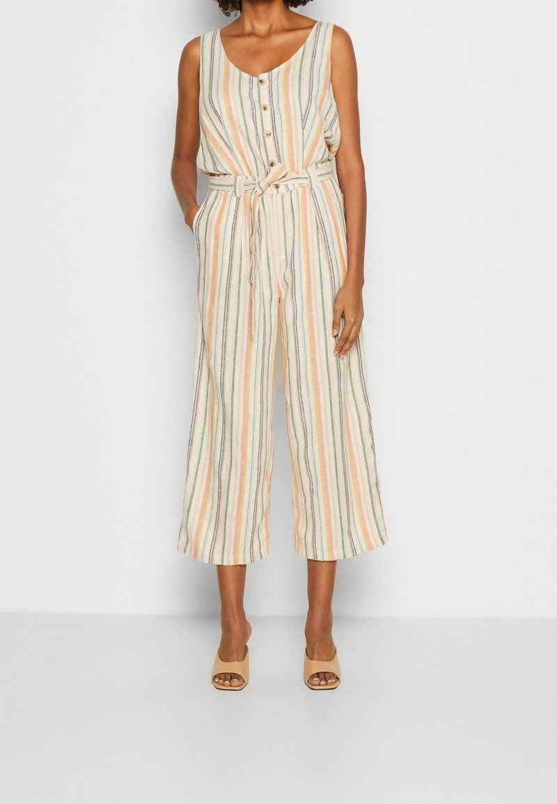 Femme portant une combinaison rayée sans manches avec un devant boutonné, une taille ceinturée, des jambes larges et des talons ouverts beige sur fond blanc.