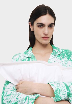 Camicetta a motivi verdi e bianchi con colletto; modello che tiene un cuscino bianco. La texture appare morbida, con pieghe visibili nel tessuto.