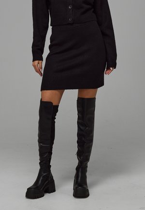 Femme portant une jupe en maille noire et un haut assorti, assorties à des bottes noires montantes épaisses, debout sur un sol et un arrière-plan gris.