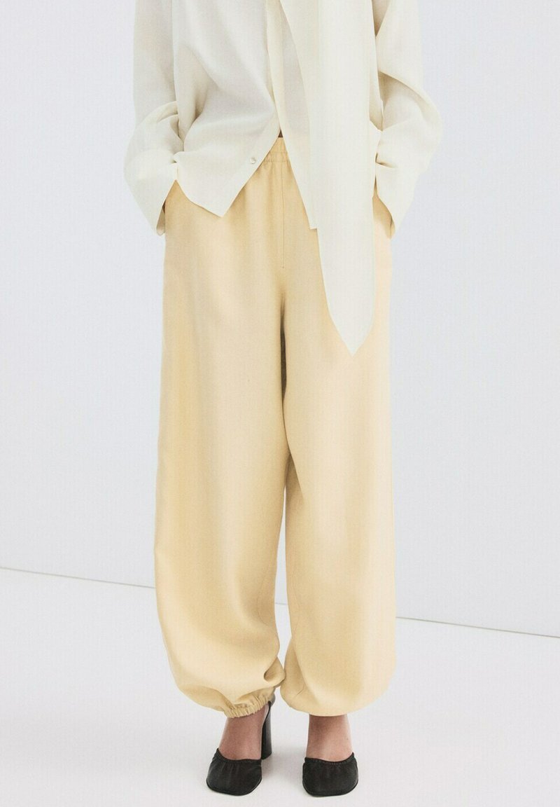 Pantalon ample beige avec chevilles élastiques porté avec des chaussures noires à talons et un haut crème à manches longues, les mains dans les poches.
