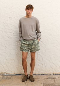 Hellgrauer, langärmliger Pullover, grüne gemusterte Badeshorts und braune Sandalen mit Riemchen, getragen von einem Model vor einer strukturierten weißen Wand.