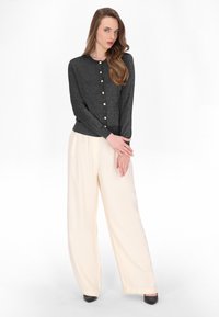 Cardigan nero scintillante con scollo tondo e chiusura a bottoni, abbinato a pantaloni crema a gamba larga. La modella indossa tacchi neri.