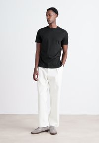 Camiseta de algodón negra combinada con pantalones blancos de cintura alta. El modelo está de pie sobre una superficie clara, llevando zapatos grises, con las manos en los bolsillos.