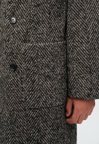 Detail ruky vedle hnědo-černého kabátu s herringbone vzorem, černými knoflíky a velkou čtvercovou kapsou.