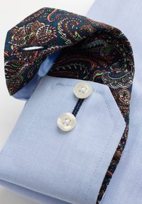 Lichtblauw overhemdmanchet met twee witte knopen en een voering van stof met een paisley-patroon in donkerblauwe, rode en groene tinten.