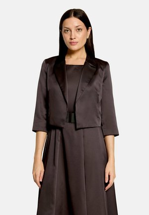 Femme aux longs cheveux foncés porte un blazer court marron foncé à manches trois quarts sur une robe assortie ceinturée.