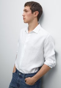 Weißes Leinenhemd mit Button-Down-Kragen und aufgerollten Ärmeln, kombiniert mit dunkelblauen Jeans, die einen lässigen Look kreieren.