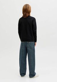 Vállig érő világosbarna hajú személy hátulról nézve, fekete pulóvert, széles szárú kék farmert és fehér tornacipőt visel.