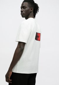 Λευκό βαμβακερό t-shirt με στρογγυλή λαιμόκοψη. Κόκκινο γραφικό έμπλαστρο στην πλάτη με τη λέξη "HUGO." Το μοντέλο στέκεται πλάγια, φορώντας μαύρο σορτς.