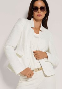 Witte getextureerde blazer met subtiele versieringen, met een reverskraag en één knoopsluiting. Gecombineerd met een witte top en riem.