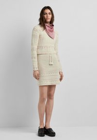 Donna che indossa un completo di maglione e gonna in crochet color crema, sciarpa rosa al collo e zoccoli neri, in piedi contro un muro grigio uniforme.