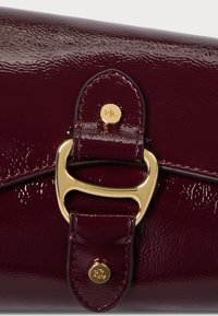 Közelkép egy fényes sötét burgundi bőr táskával, arany színű fém csattal és két arany szegeccsel, amelyeken "RL" van gravírozva.