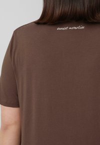 Bruine T-shirt met korte mouwen, gemaakt van zachte stof, met "zonnenuntergang herinneringen" in licht borduurwerk aan de achterkant van de halslijn.