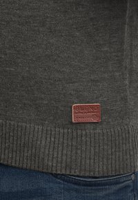 Sötétszürke kötött pulóver, bordás aljjal és egy barna bőr címkével, amelyen a "BLEND" és a "GLOBAL JEANSMAKERS" felirat van gravírozva.