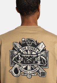 Beige t-shirt med et sort grafisk design, der viser en hammer, skateboarddele og stammer, mærket "THE DOERS" og "ELEMENT X TIMBER!"