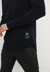 Tmavě námořnická košile s dlouhým rukávem a kulatým lemem, se znakem v malém provedení a mírně texturovanou tkaninou. Nositá přes černé kalhoty.