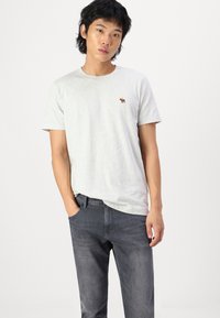 Grå t-skjorte med korte ermer som har et lite brunt dyrelogo. Bæreren har på seg mørkegrå jeans med hendene i lommene. Enkel bakgrunn.