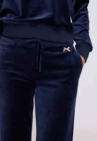 Navy blauwe velours trainingspak met een geribde tailleband, trekkoord-detail en een kleine beige strikaccent op de zijzak.